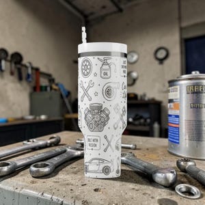 Op de afbeelding: Een witte beker met een zwart-wit ontwerp met autothema, inclusief moersleutels, een automotor en de tekst "Built with Passion". De beker heeft een wit deksel en een rietje. De achtergrond bevat gereedschap en een oliebuss.
