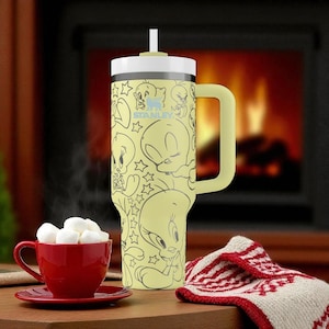 May include: A yellow Stanley tumbler featuring a cartoon character design, a handle, and a straw. The tumbler is next to a red teacup filled with marshmallows and a red and white patterned cloth. The background includes a fireplace.