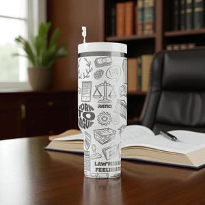 May include: White tumbler with a black and white law-themed design. The tumbler features illustrations of scales, money, a gavel, and the words "Born to Argue" and "Justice". It has a white lid and a straw.