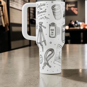 K&ouml;nnte beinhalten: Wei&szlig;er Reisebecher mit gro&szlig;em Griff, verziert mit schwarzen Strichzeichnungen von Gegenst&auml;nden aus der Fotografie, darunter ein Kameraobjektiv, eine Filmrolle und ein Stativ. Der Text "Capture The Moment" ist ebenfalls enthalten.