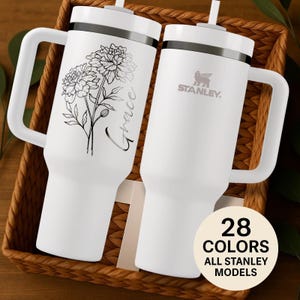 May include: Two white stainless steel tumblers with handles and straws. One tumbler features a black line art floral illustration and the script word Grace. The other tumbler displays the Stanley logo. A circular badge reads 28 COLORS ALL STANLEY MODELS.