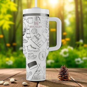 Op de afbeelding: Een witte Stanley-beker met handvat en rietje, voorzien van een zwart lijnpatroon met outdoor-iconen zoals bergen, dennenbomen, een kompas, een wandelschoen, een mok en bordjes met de tekst hiking en fishing op de buitenkant.