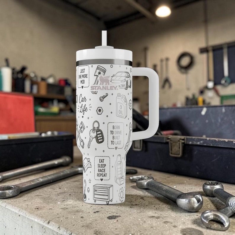 Op de afbeelding: Een witte Stanley-beker met een handvat en een rietje, versierd met zwarte lijntekeningen van auto-gerelateerde iconen en tekst. De tekst bevat "Car Life", "Eat Sleep Race Repeat" en "Born to Drive Built to Last."
