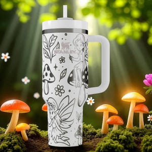 May include: White Stanley travel mug with a handle, decorated with a fairy and mushroom design. The mug has a white lid and straw. The design features black and white illustrations of fairies, mushrooms, and floral elements.