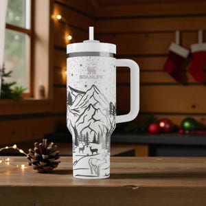 K&ouml;nnte beinhalten: Wei&szlig;er Stanley-Becher mit einem schwarz-wei&szlig;en Berg- und Hirschdesign. Der Becher hat einen wei&szlig;en Griff und Deckel mit Strohhalm. Das Stanley-Logo ist sichtbar. F&uuml;r kalte Getr&auml;nke konzipiert.