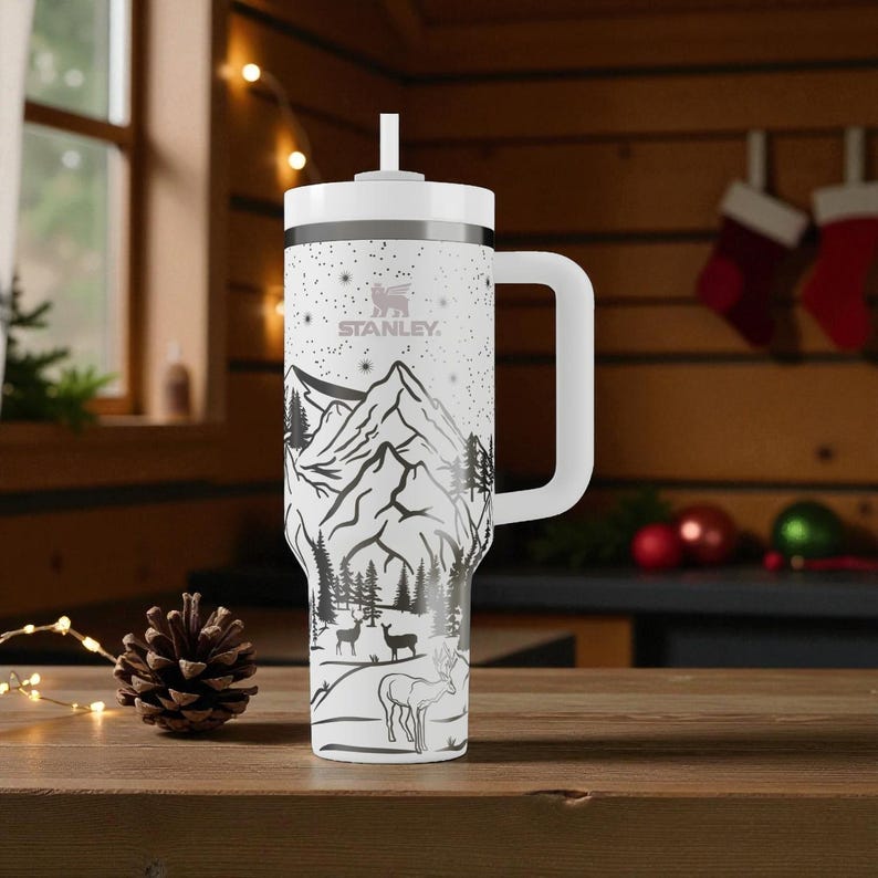 K&ouml;nnte beinhalten: Wei&szlig;er Stanley-Becher mit einem schwarz-wei&szlig;en Berg- und Hirschdesign. Der Becher hat einen wei&szlig;en Griff und Deckel mit Strohhalm. Das Stanley-Logo ist sichtbar. F&uuml;r kalte Getr&auml;nke konzipiert.