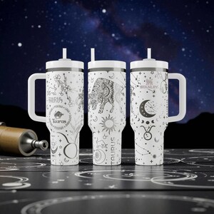 May include: Three white Stanley tumblers with handles and straws, featuring black line art of the Taurus zodiac sign, bulls, moons, stars, and floral patterns. The designs include the word Taurus and various astrological symbols printed on the exterior.