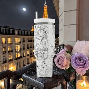 May include: White tumbler with a black and white anime design featuring a character with long hair and a sailor uniform. The tumbler has a white lid and a straw. The background includes a night scene with the Eiffel Tower and a full moon.
