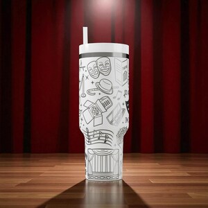May include: A white tumbler with a black and white theater-themed design, including comedy and tragedy masks, musical notes, and a stage curtain. The tumbler has a white lid and a straw. The background features a red curtain and a wooden floor.