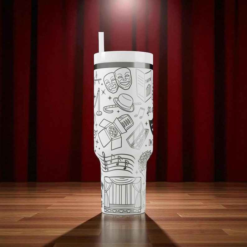 May include: A white tumbler with a black and white theater-themed design, including comedy and tragedy masks, musical notes, and a stage curtain. The tumbler has a white lid and a straw. The background features a red curtain and a wooden floor.
