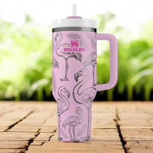 May include: A light pink Stanley tumbler with a handle, a white lid, and a straw. The exterior features a repeating pattern of dark grey line-art flamingos and feathers. The Stanley logo is printed in dark pink near the top of the cup.