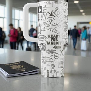May include: A white travel mug with a large handle, decorated with black and white travel-themed illustrations, such as aeroplanes and passport stamps. The text "READY FOR TAKEOFF" is visible. A passport is placed next to the mug.
