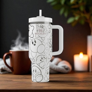 May include: A white Stanley tumbler with a handle and a straw, featuring a black and white line art pattern of various Kirby characters and stars. The Stanley logo is printed in black on the upper portion of the cylindrical insulated cup.