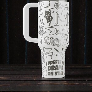 May include: White travel mug with a large handle, decorated with black theater-themed illustrations. The design includes a ballerina, piano, and the text "I prefer my drama on stage."