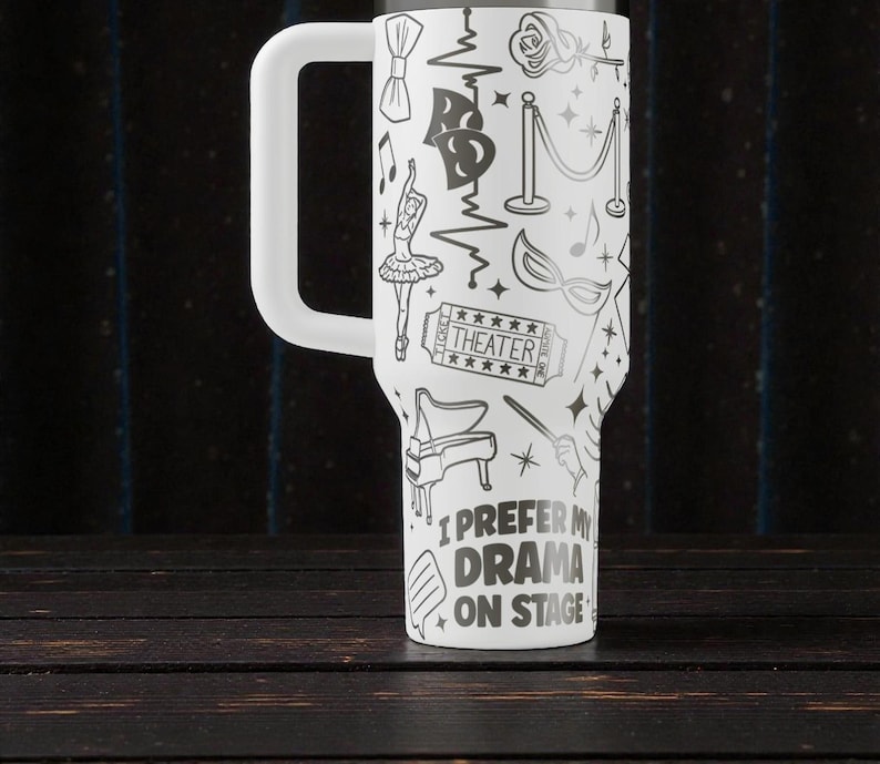 May include: White travel mug with a large handle, decorated with black theater-themed illustrations. The design includes a ballerina, piano, and the text "I prefer my drama on stage."