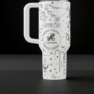 K&ouml;nnte beinhalten: Wei&szlig;er Becher mit einem gro&szlig;en wei&szlig;en Griff, mit einem Steinbock-Tierkreiszeichen-Design. Das Design enth&auml;lt das Wort "Steinbock", eine Ziegenillustration und andere Tierkreissymbole. Der Becher ist mit Sternen, Monden und floralen Elementen verziert.