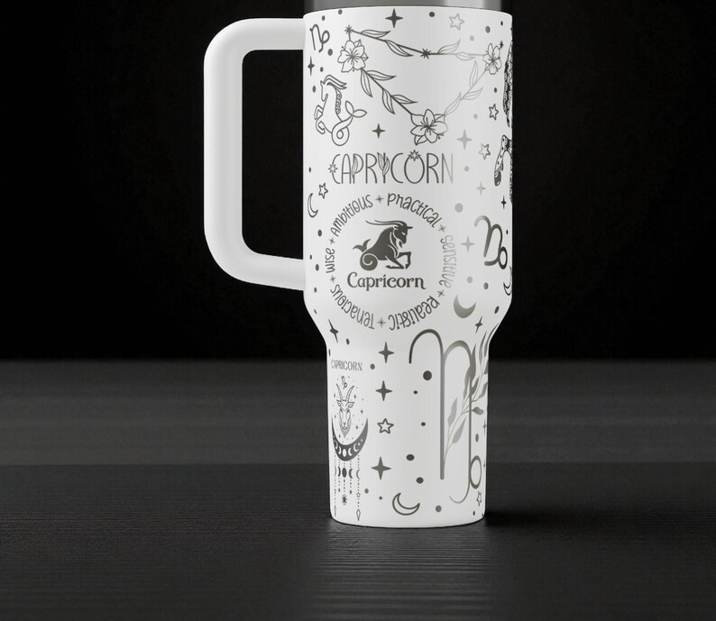 K&ouml;nnte beinhalten: Wei&szlig;er Becher mit einem gro&szlig;en wei&szlig;en Griff, mit einem Steinbock-Tierkreiszeichen-Design. Das Design enth&auml;lt das Wort "Steinbock", eine Ziegenillustration und andere Tierkreissymbole. Der Becher ist mit Sternen, Monden und floralen Elementen verziert.