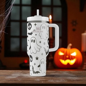 May include: A white Stanley tumbler with a handle, lid, and straw. The tumbler is decorated with black Halloween-themed graphics, including ghosts, bats, skulls, and stars. The background includes a carved pumpkin and a lit candle.