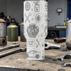 Op de afbeelding: Een witte tumbler met een autothema, versierd met auto-gerelateerde illustraties. Het ontwerp bevat afbeeldingen van automotoren, zuigers en sleutels. De tekst "Built with Passion" en "Just One More Mod" zijn ook zichtbaar.