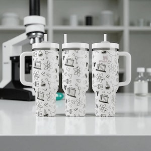 May include: Three white Stanley tumblers with black science-themed illustrations, including beakers and atoms. Each tumbler has a white handle, lid, and straw. The tumblers are on a white surface, with a microscope in the background.