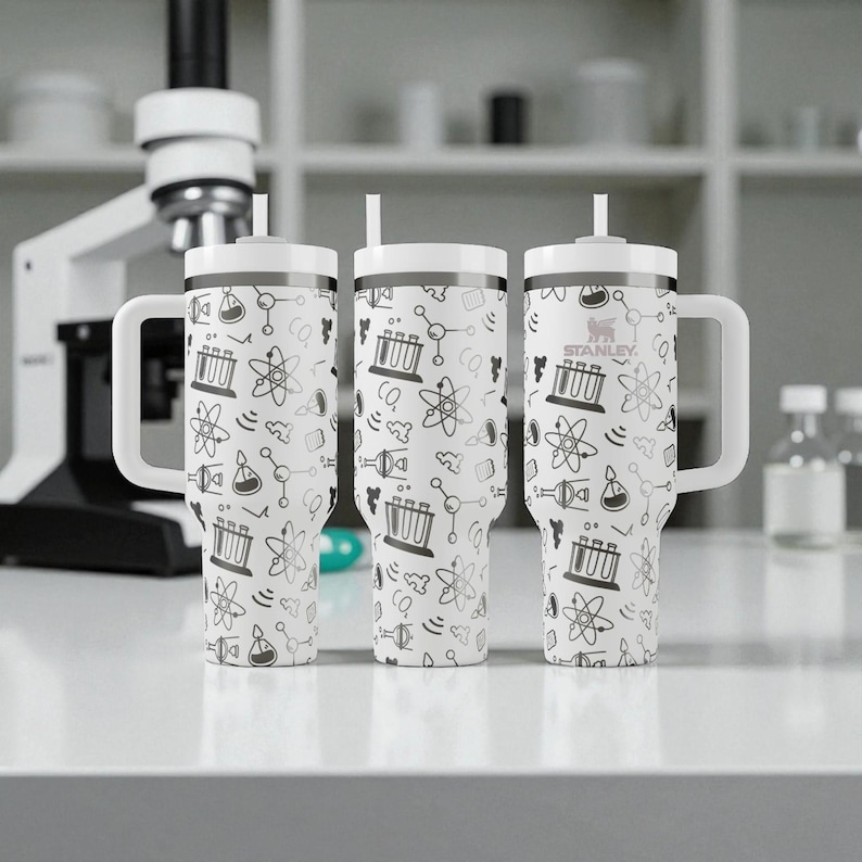 May include: Three white Stanley tumblers with black science-themed illustrations, including beakers and atoms. Each tumbler has a white handle, lid, and straw. The tumblers are on a white surface, with a microscope in the background.