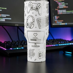 May include: White tumbler with black line art of gaming-related icons, including a controller, headphones, keyboard, and a computer screen with a loading bar. The text "Stay Wired" and "GG" are also featured.