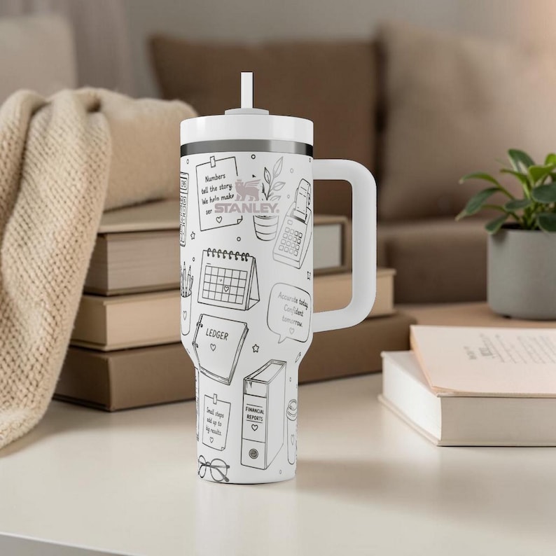Puede incluir: Vaso Stanley blanco con una ilustraci&oacute;n en blanco y negro de elementos relacionados con la contabilidad, incluyendo una calculadora, un calendario y un libro mayor. El vaso tiene un asa y una tapa blancas con una pajita. El texto en el vaso incluye "Numbers tell the story" y "STANLEY."