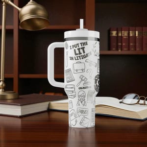 May include: White travel mug with a handle and a straw, featuring a black and white doodle design. The mug has the text "I PUT THE LIT IN LITIGATE" and includes illustrations of legal symbols and objects. The mug is on a wooden desk.