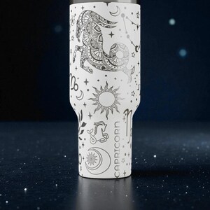 K&ouml;nnte beinhalten: Wei&szlig;er Becher mit einem schwarzen Capricorn-Tierkreiszeichen-Design, einschlie&szlig;lich Sterne, Monde und Sonnen. Das Wort "CAPRICORN" ist vertikal aufgedruckt. Der Becher hat einen Edelstahlrand und einen wei&szlig;en K&ouml;rper.