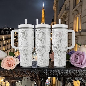 May include: Three white insulated tumblers with black line art of anime characters, each with a handle and a straw. The tumblers are on a black metal railing with flowers in the foreground and the Eiffel Tower in the background.