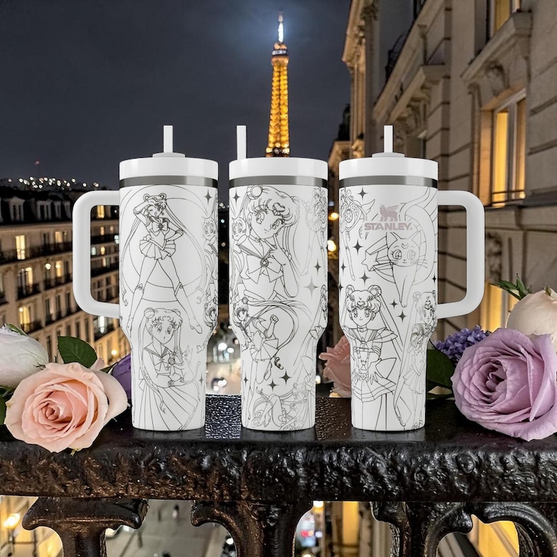 May include: Three white insulated tumblers with black line art of anime characters, each with a handle and a straw. The tumblers are on a black metal railing with flowers in the foreground and the Eiffel Tower in the background.