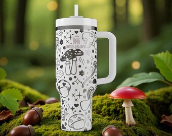May include: A white Stanley tumbler with a handle and straw, featuring a black line-art pattern of mushrooms, hearts, flowers, and stars. The brand name Stanley is printed in black near the top of the cup, which has a white lid and a white straw.