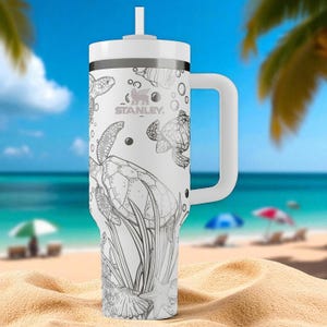 May include: White Stanley travel mug featuring a black and white sea turtle illustration. The mug has a white handle and lid with a straw. The background shows a beach scene with sand, sea, and palm trees.