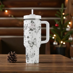 Może przedstawiać: Biały kubek Stanley z czarnym wzorem motyli i gwiazd. Kubek ma biały uchwyt i pokrywkę ze słomką. Słowo "STANLEY" jest nadrukowane z boku. Dekoracyjny przedmiot na napoje.