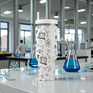 May include: A white insulated tumbler with a black and gray science-themed pattern, including beakers, atoms, and molecules. The tumbler has a white lid and a straw. The background shows a laboratory setting with glassware and scientists.