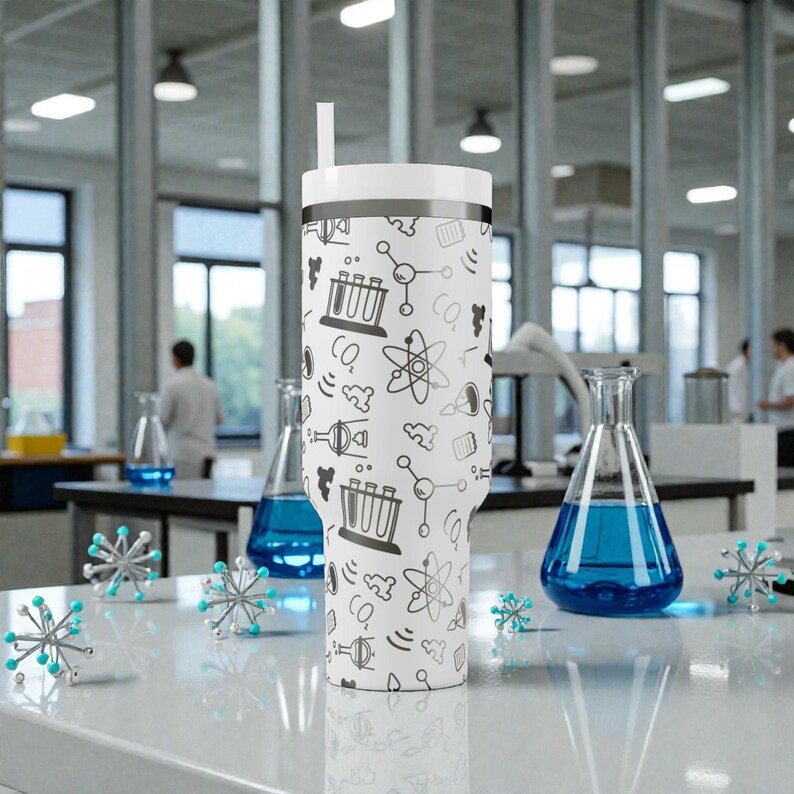 May include: A white insulated tumbler with a black and gray science-themed pattern, including beakers, atoms, and molecules. The tumbler has a white lid and a straw. The background shows a laboratory setting with glassware and scientists.