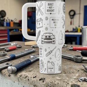 Op de afbeelding: Een witte reismok met een grote handgreep, versierd met een herhalend patroon van zwarte illustraties met een mechanisch thema. De mok heeft de tekst "Built - Not - Bought" en "Mechanic Mode On". Verschillende gereedschappen liggen rond de mok.