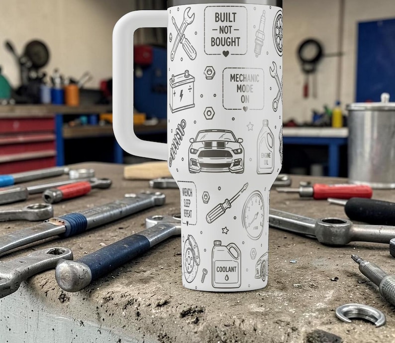 Op de afbeelding: Een witte reismok met een grote handgreep, versierd met een herhalend patroon van zwarte illustraties met een mechanisch thema. De mok heeft de tekst "Built - Not - Bought" en "Mechanic Mode On". Verschillende gereedschappen liggen rond de mok.