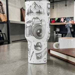 K&ouml;nnte beinhalten: Ein wei&szlig;er Reisebecher mit einer schwarz-wei&szlig;en Illustration einer Kamera, Bergen und anderen Elementen der Fotografie. Der Becher tr&auml;gt den Text "Focus On The Good" und steht auf einer grauen Oberfl&auml;che.