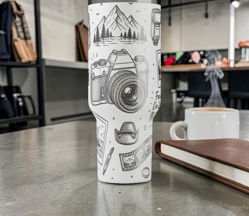 K&ouml;nnte beinhalten: Ein wei&szlig;er Reisebecher mit einer schwarz-wei&szlig;en Illustration einer Kamera, Bergen und anderen Elementen der Fotografie. Der Becher tr&auml;gt den Text "Focus On The Good" und steht auf einer grauen Oberfl&auml;che.