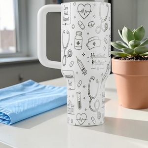 Puede incluir: Un vaso blanco con un asa blanca grande, decorado con ilustraciones de tem&aacute;tica m&eacute;dica en negro. El vaso presenta estetoscopios, pastillas y el texto "Heartbeat Matters". Un pa&ntilde;o azul y una suculenta en maceta est&aacute;n en el fondo.