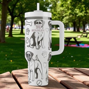 May include: White Stanley tumbler with a black and gray dog pattern, paw prints, and bone illustrations. Features a white handle, lid, and straw. The tumbler is designed for cold beverages and is ideal for outdoor use.