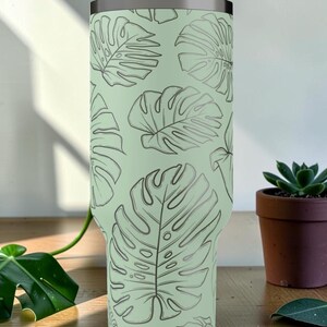 Puede incluir: Un vaso verde claro con un patr&oacute;n repetido de hojas de monstera grises. El vaso de acero inoxidable tiene un borde plateado. Una peque&ntilde;a suculenta en maceta se encuentra sobre una superficie de madera.