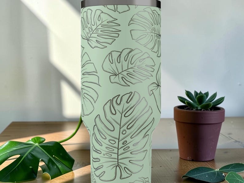 Puede incluir: Un vaso verde claro con un patr&oacute;n repetido de hojas de monstera grises. El vaso de acero inoxidable tiene un borde plateado. Una peque&ntilde;a suculenta en maceta se encuentra sobre una superficie de madera.
