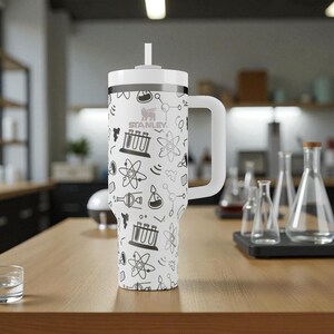 May include: A white Stanley tumbler with a handle and straw, adorned with a black and gray science-themed pattern. The design includes beakers, atoms, and other lab-related illustrations. The tumbler is on a wooden surface.