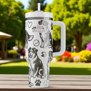 May include: A white Stanley tumbler with a handle and a straw, featuring a repeating black graphic print of Australian Shepherd dogs, paw prints, hearts, and tennis balls. The Stanley logo is printed in black on the upper portion of the cup.