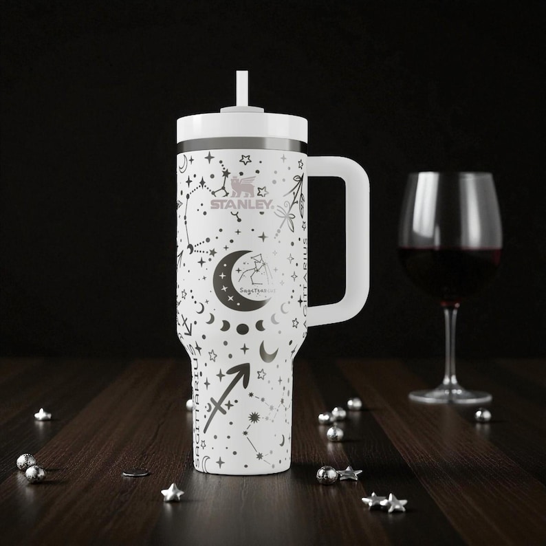 Op de afbeelding: Witte Stanley-beker met een handvat, versierd met een zwart sterrenbeeldontwerp, het Boogschutter-symbool en het Stanley-logo. Het heeft een wit deksel en een rietje. Een wijnglas met rode wijn staat ernaast.