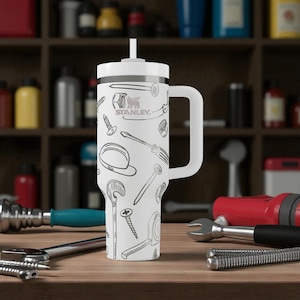 Puede incluir: Un vaso Stanley blanco con asa y pajita, que presenta un patrón de líneas negras de varias herramientas de ferretería, incluyendo tornillos, clavos, tuercas, un destornillador y una cinta métrica impresos en la superficie exterior.