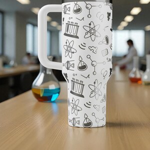 May include: A white insulated tumbler with a large white handle, covered in a repeating pattern of science-themed doodles in black. The doodles include atoms, beakers, and test tubes. The tumbler is sitting on a wooden table.