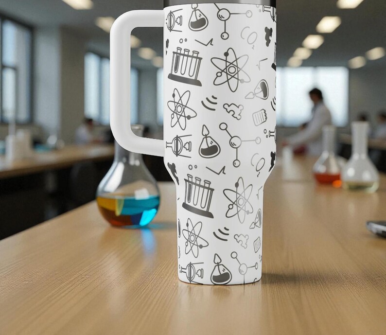 May include: A white insulated tumbler with a large white handle, covered in a repeating pattern of science-themed doodles in black. The doodles include atoms, beakers, and test tubes. The tumbler is sitting on a wooden table.